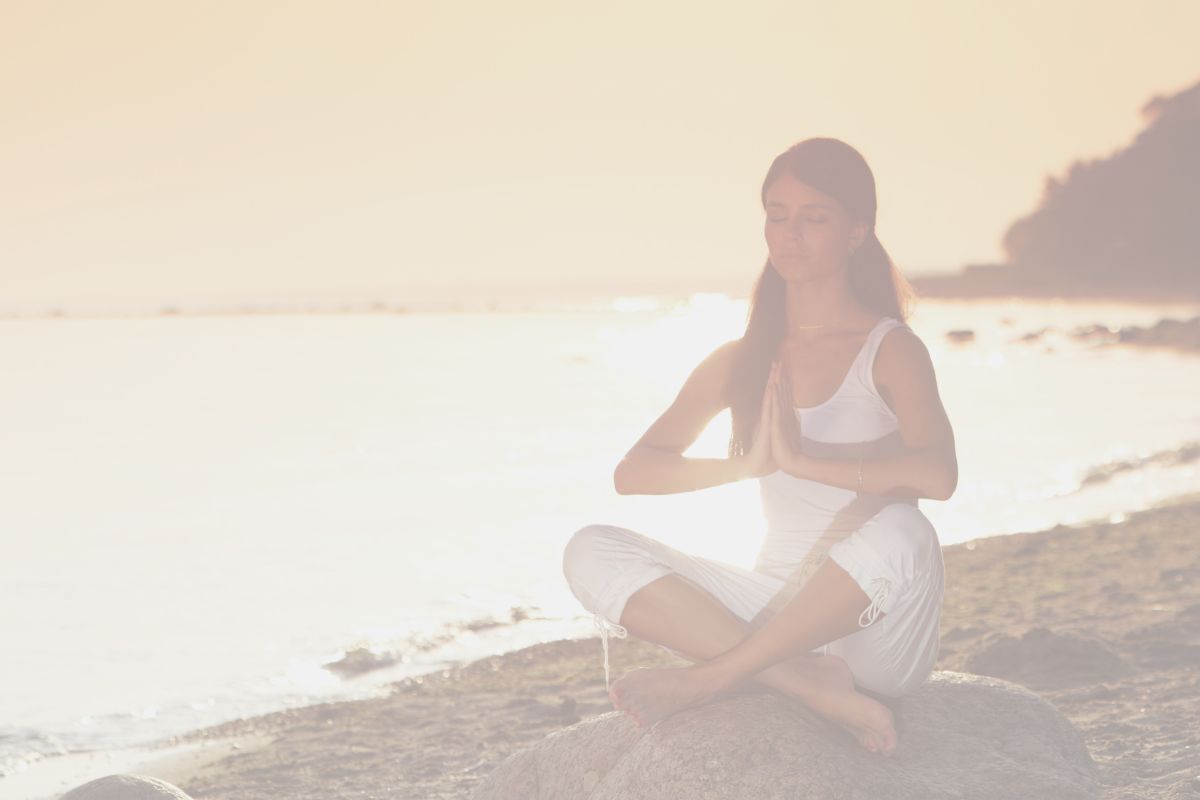 woman in white yoga clothes meditating on the beach at sunrise showing time of day as one of the themes of yoga classes