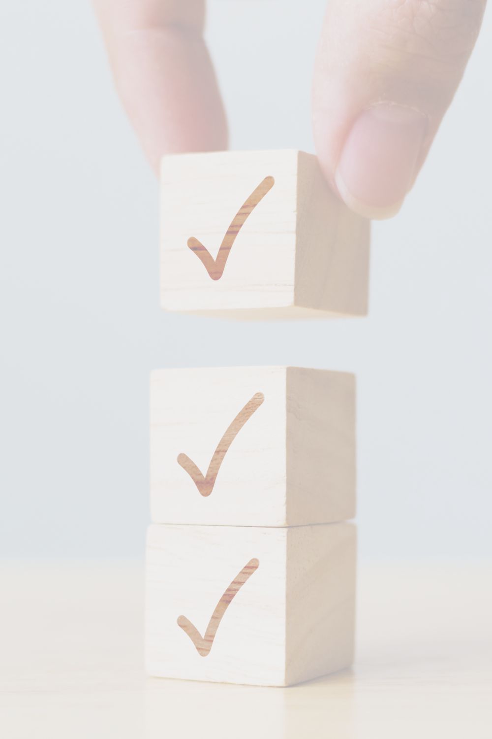 Two wooden cubes each with a check mark are stacked on top of each other. A third identical cube is being placed on top, held between someone's thumb and index finger.