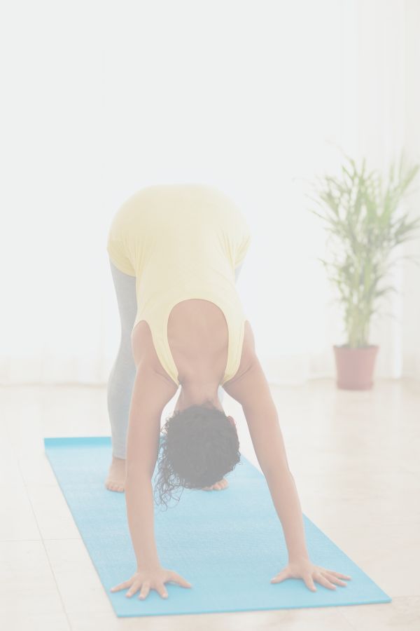 A woman in grey leggings and a yeallow tanktop is practicing Downward Facing Dog on a blue yoga mat indoors in the sunshine.