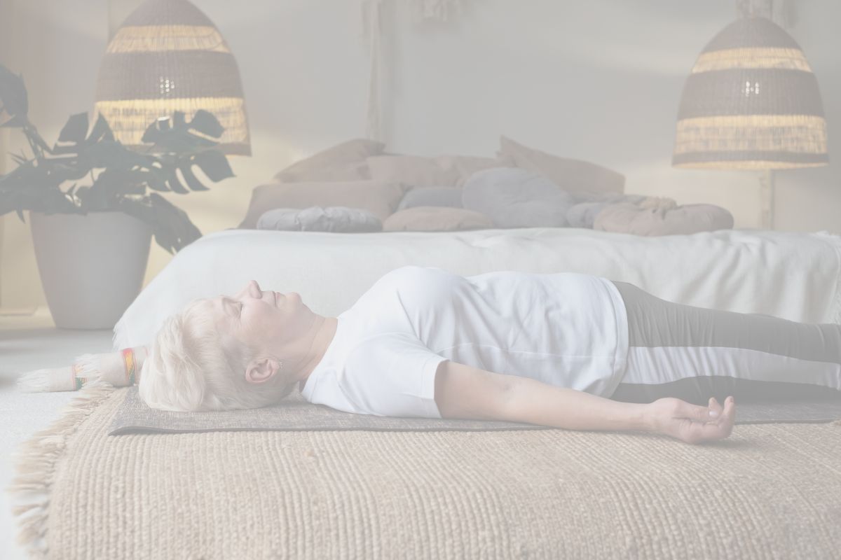 A woman in a white tee-shirt is lying down on a yoga mat in front of a bed.
