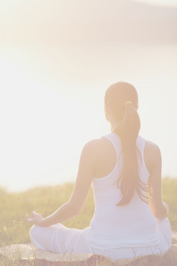 A woman in white yoga clothes is meditating on a rock. She is facing away from the camera towards the evening sun setting over a grassy field.