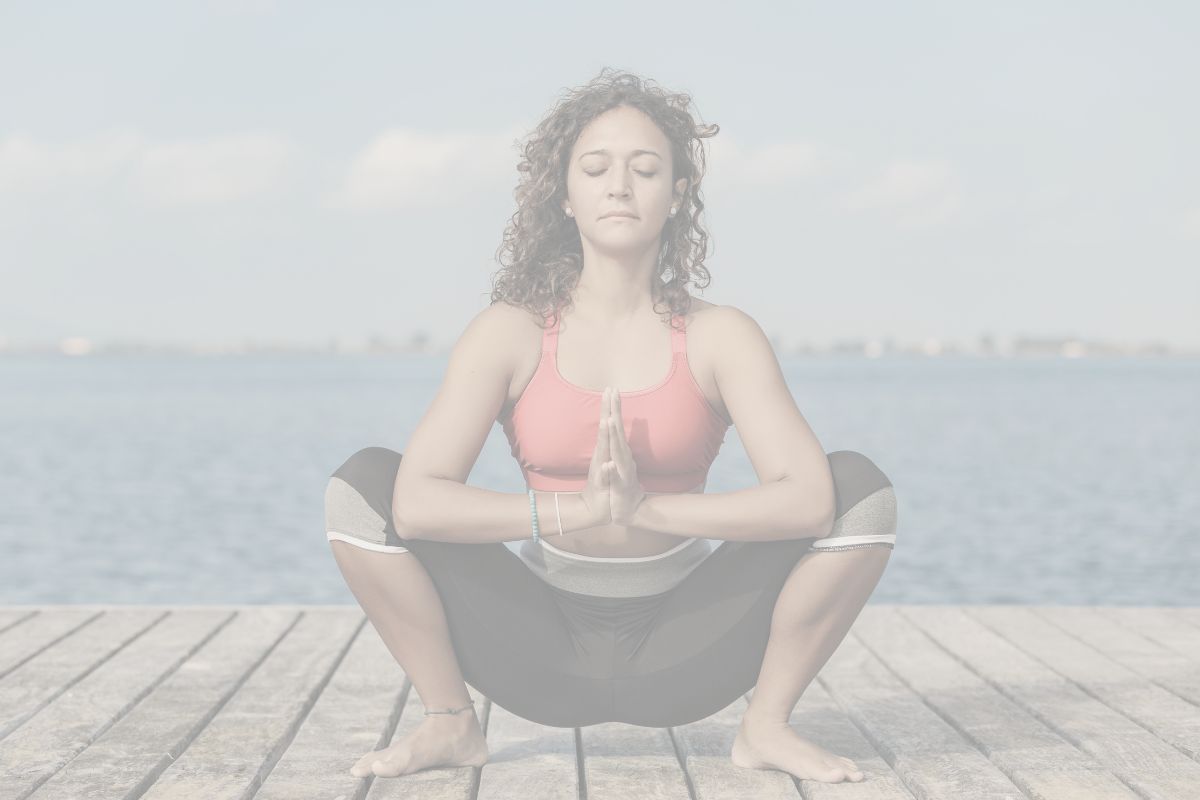 A woman on a deck by the ocean is in a deep yoga squat pose with her hands in prayer.