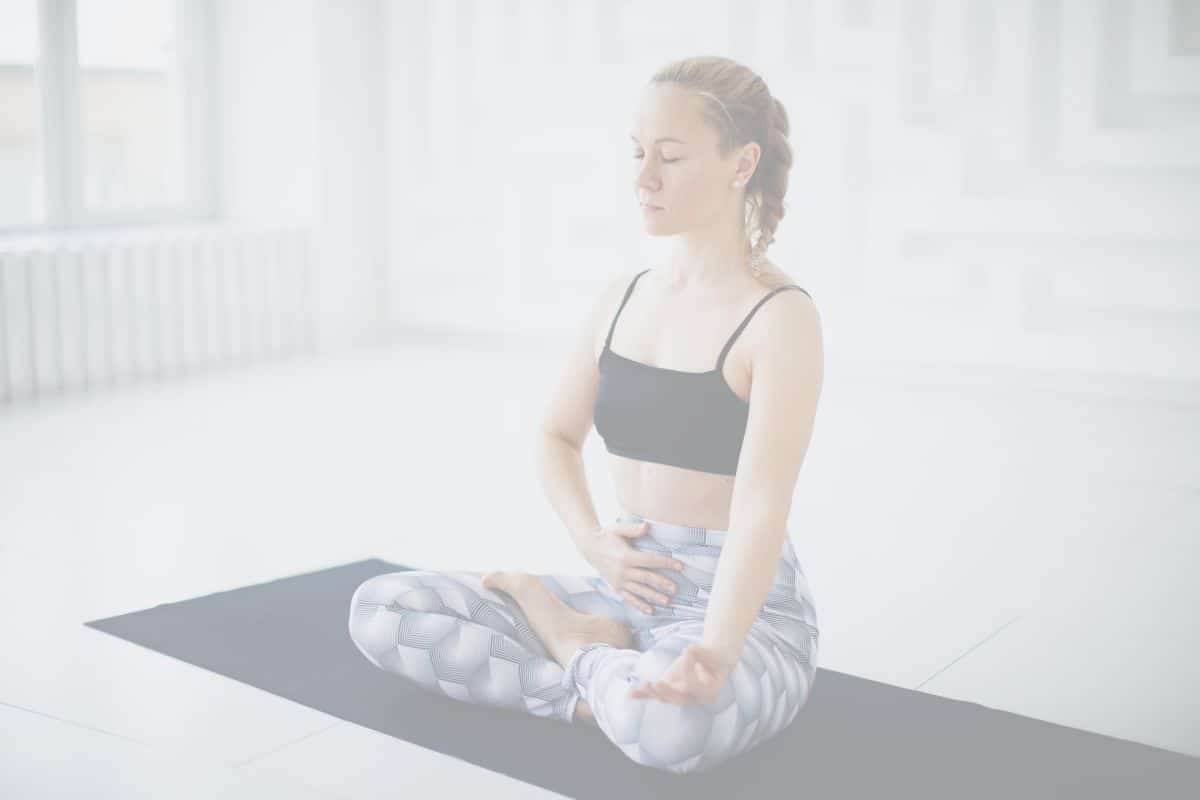 A young woman is sitting on a yoga mat with her eyes closed and one hand on her belly, practicing Belly Breath, or Adham Pranayama