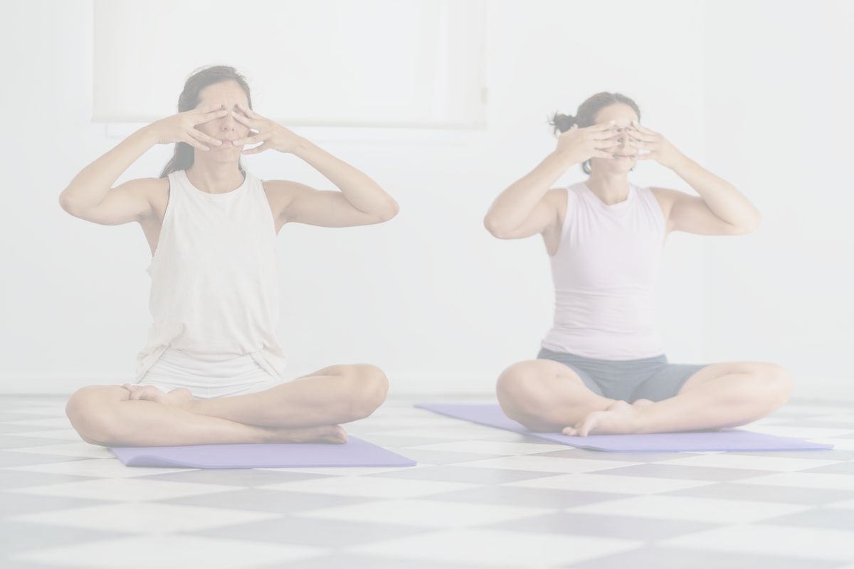 Two women are sitting cross-legges with they fingers over their eyes, demonstrating Humming Bee Breath, a practice suitable for yoga beginners and home yoga practitioners.