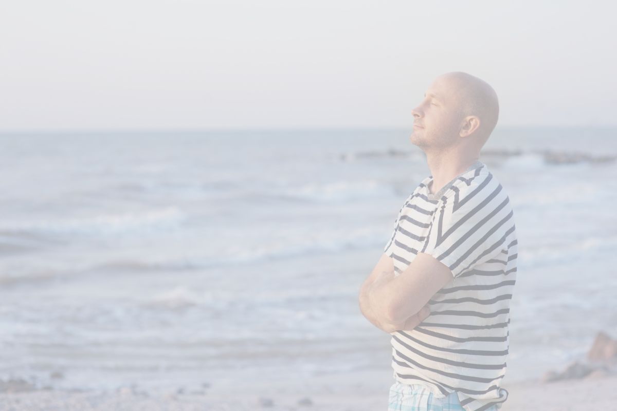 A man in a striped shirt stands next to an ocean. you can see waves rolling in, likely making the sound similar to that in Ujjayi Breath.