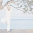 A man in loose white yoga clothes is practising tree pose underneath a tree