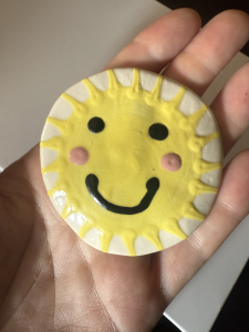 A hand holding a ceramic disc with a yellow sun painted on, with a smiley face.
